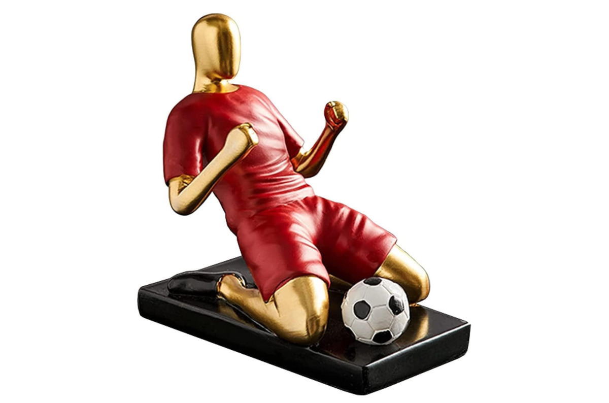 Una escultura de un futbolista con equipación de color rojo celebrando un gol con las rodillas en el suelo